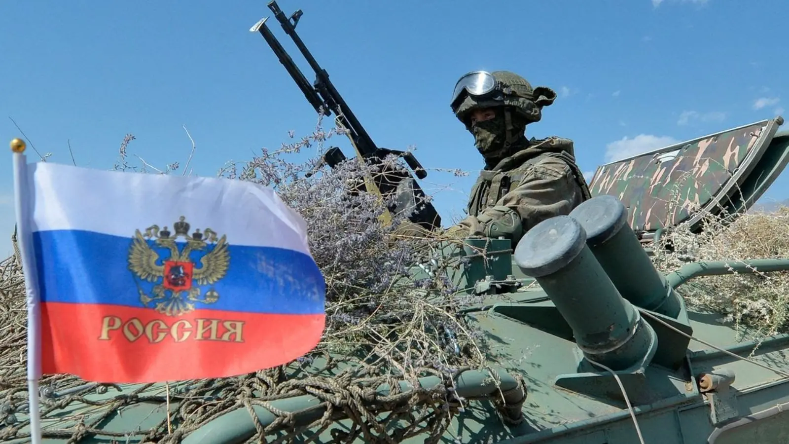 一名身穿全副武装的俄罗斯士兵，驾驶装甲车，旁边悬挂着俄罗斯国旗。