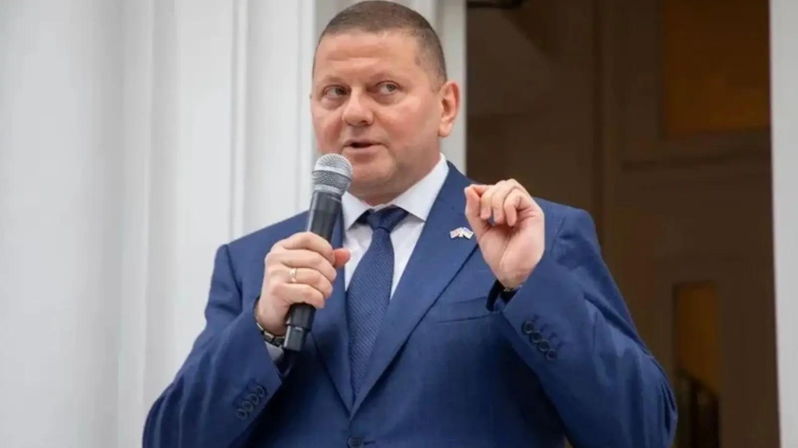 瓦列里·扎卢日尼身穿蓝色西装,对着麦克风发言并用手势示意。